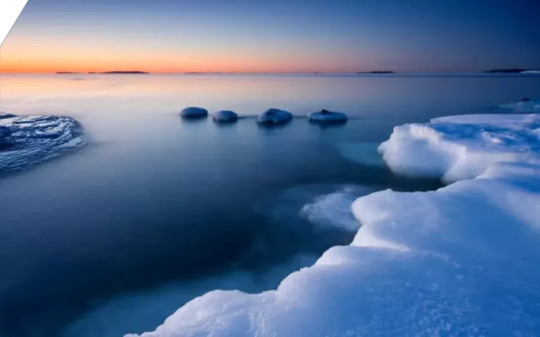 Serene winter seascape at dawn, featuring icy formations and calm, reflective water under a clear sky with shades of orange and blue.