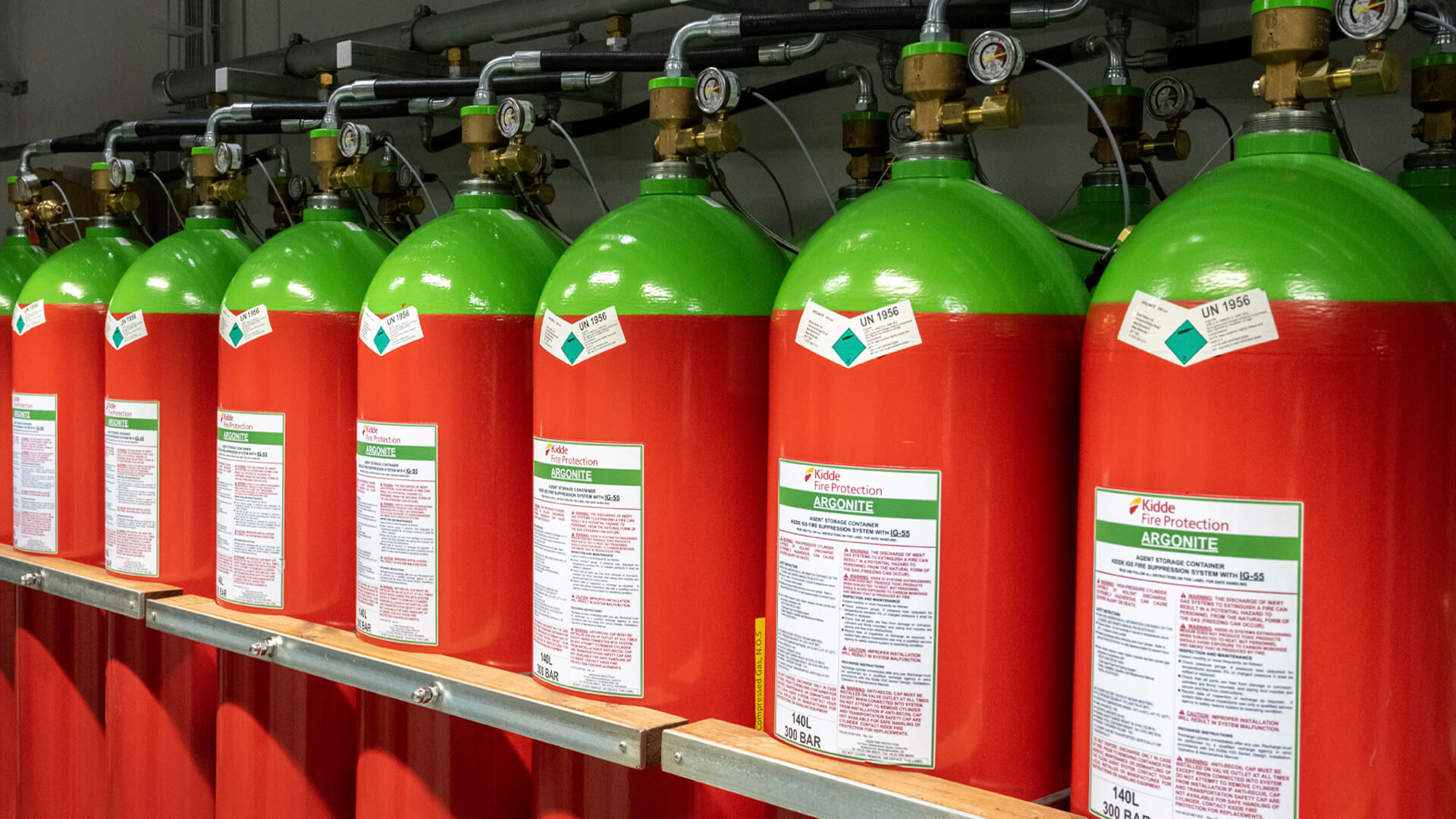 In a data center storage area, a row of red and green fire suppression gas cylinders is neatly organized, each labeled with safety warnings and details, and interconnected through a comprehensive piping system.