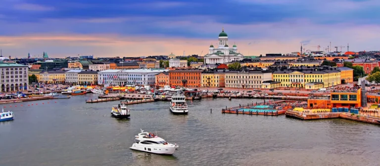 A waterfront view of Helsinki, showcasing colorful buildings and boats in the harbor, with the Helsinki Cathedral and a modern data center subtly integrated into the skyline under a cloudy sky.