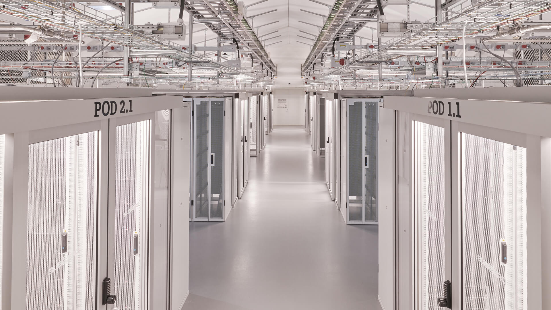 A long corridor in a modern data center showcases server racks labeled POD 2.1 and POD 1.1 on both sides. Above, the ceiling reveals an intricate network of cables and infrastructure, underscoring the complexity and efficiency of this technological hub.