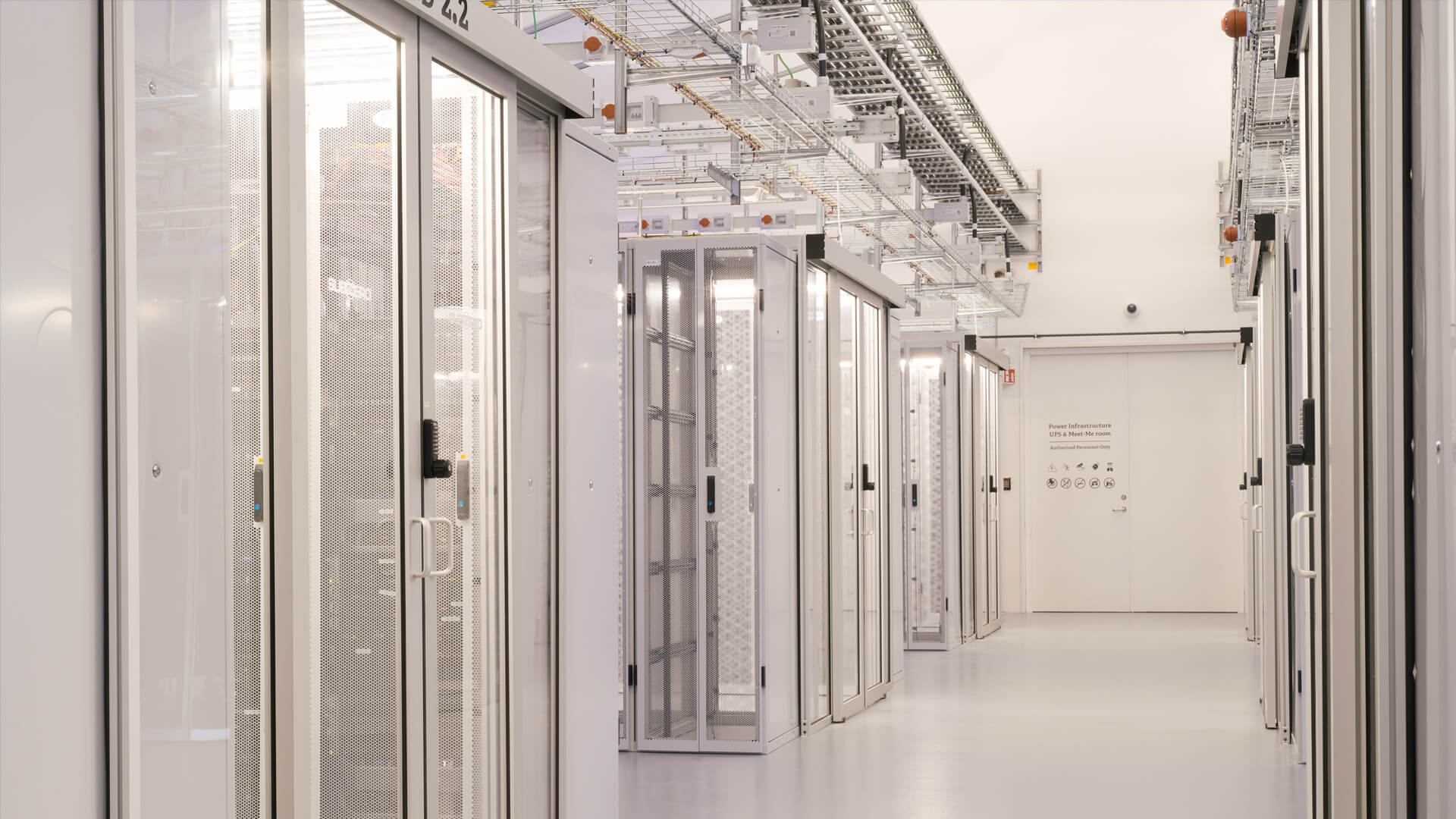 The brightly lit corridor of the data center features rows of server racks enclosed in metal cabinets, with network cables weaving visibly overhead.