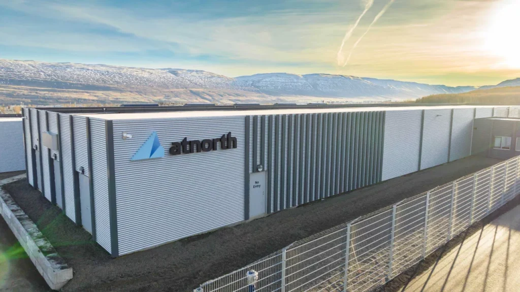 The large industrial building with metal siding, nestled behind a sturdy fence, resembles a cutting-edge data center. Snow-capped mountains stand majestically against the clear sky in the background.
