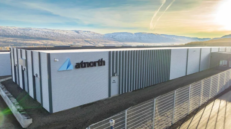 A large gray industrial building, functioning as a data center, displays the "atnorth" logo on its side. It is surrounded by a metal fence and set against a mountainous landscape under a partly cloudy sky.