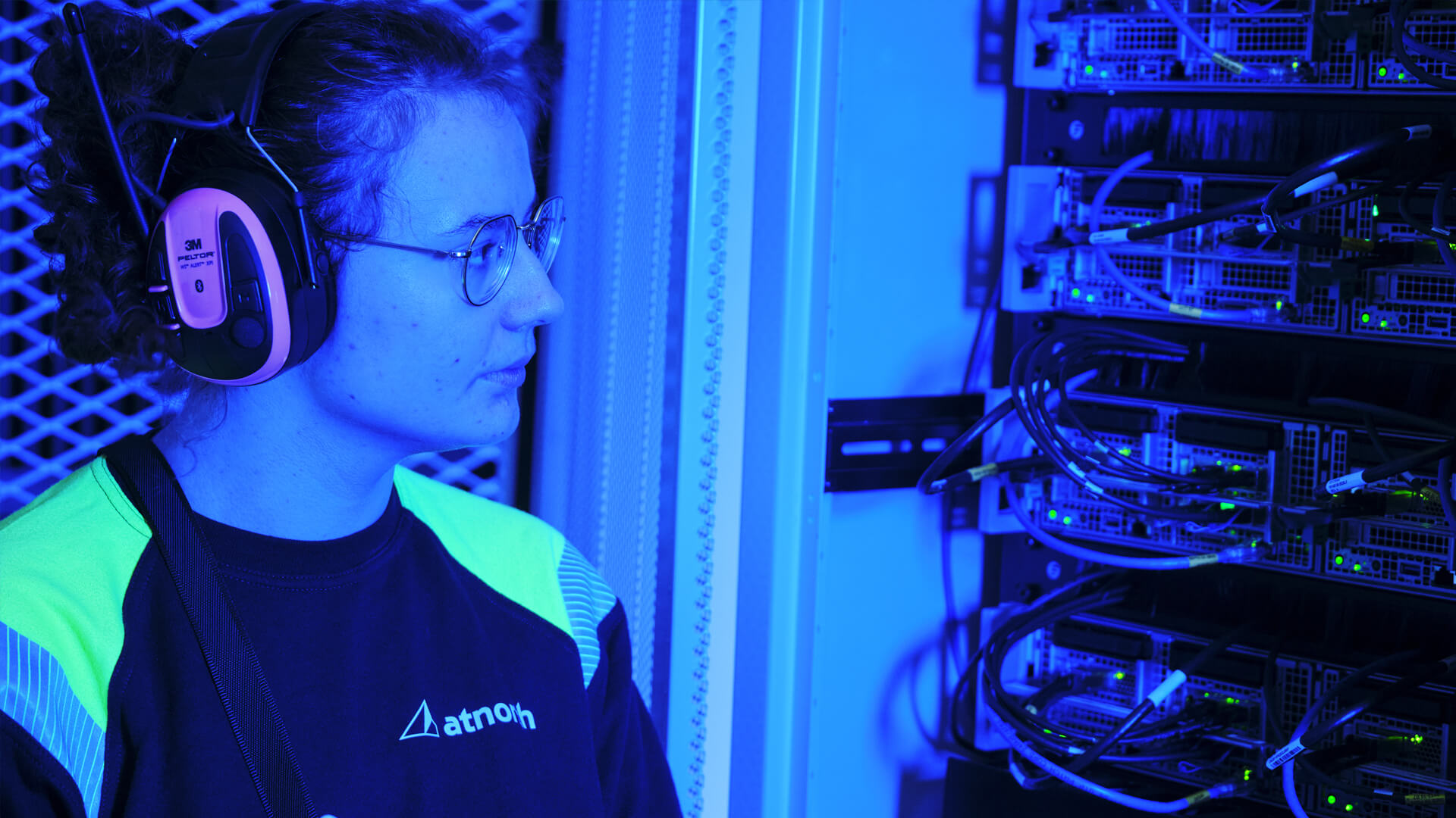 A person wearing headphones and glasses carefully monitors server equipment in a blue-lit data center.