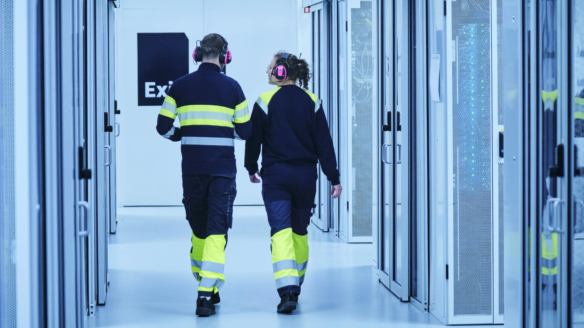 Two people in high-visibility clothing and earmuffs walk down a corridor with glass doors, heading towards an exit sign, their path lined with the hum of the data center's servers.