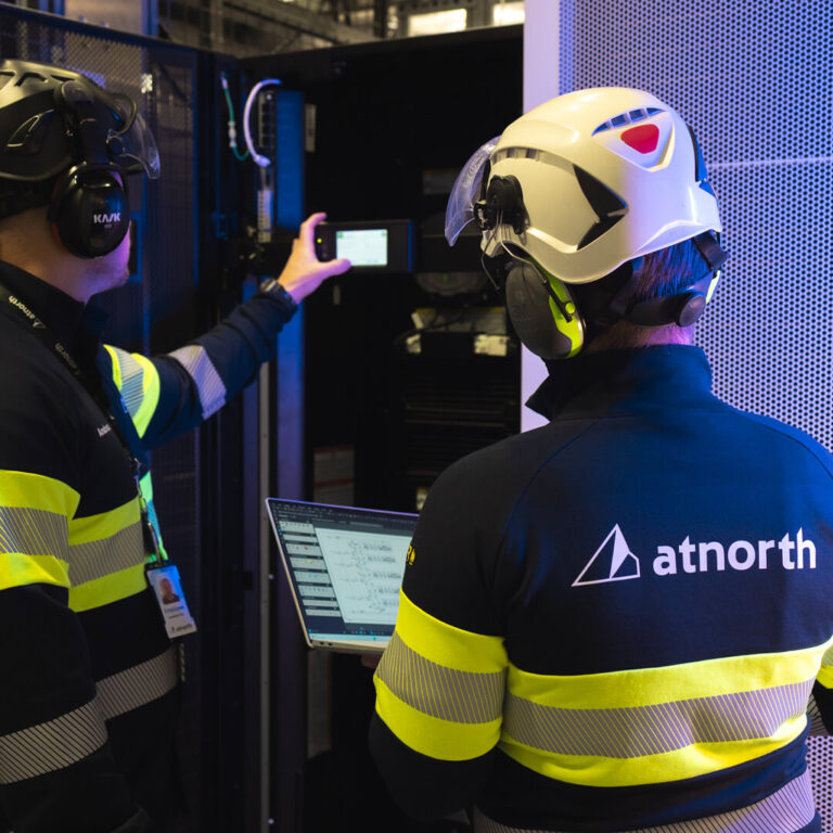 Two technicians in yellow-striped safety gear and helmets expertly manage a server rack. One holds a device, while the other uses a laptop. Emphasizing sustainable data centers, the "atnorth" logo is prominently displayed on their clothing.