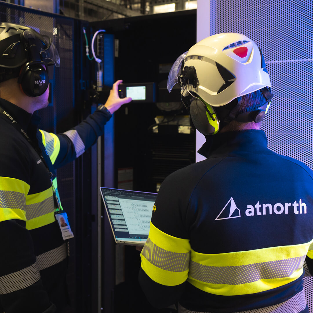 Two technicians in yellow-striped safety gear and helmets expertly manage a server rack. One holds a device, while the other uses a laptop. Emphasizing sustainable data centers, the 