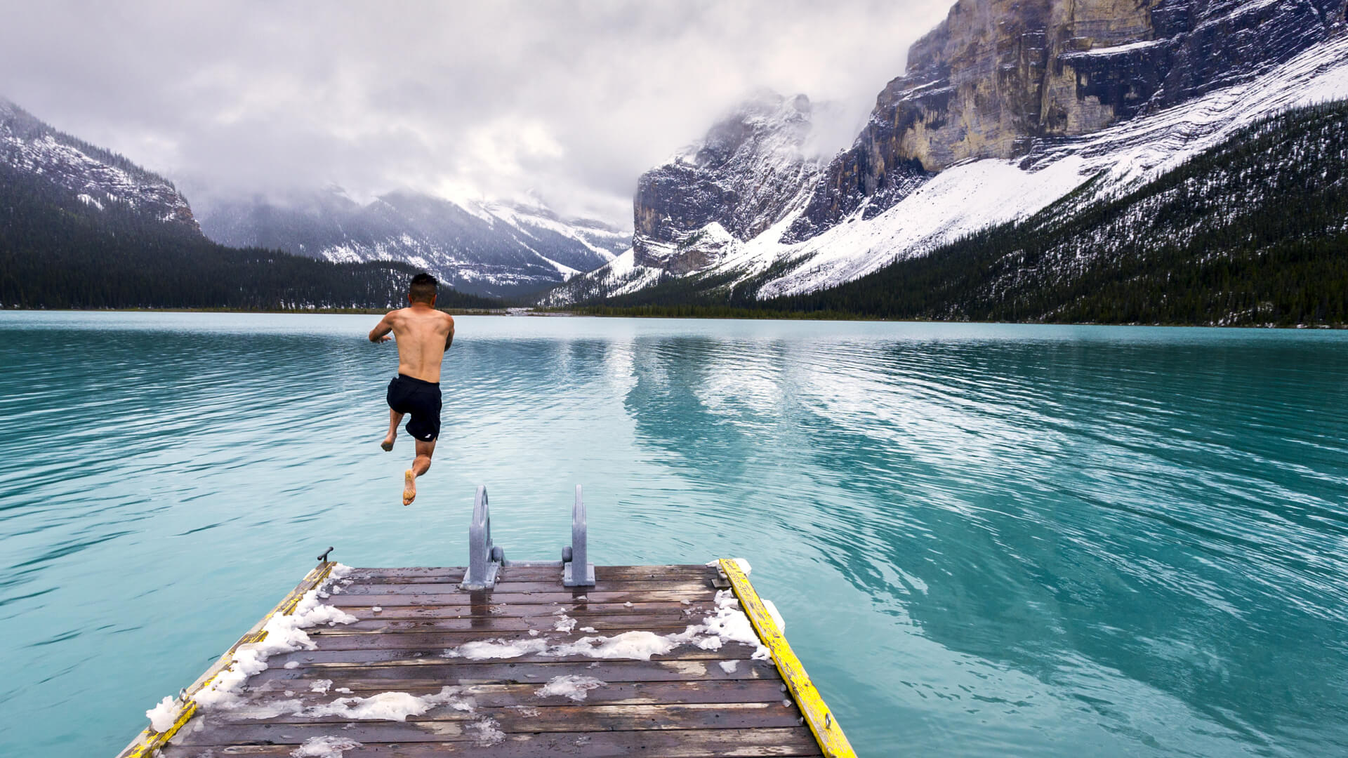 A person is leaping off a wooden dock into a turquoise lake, surrounded by snow-capped mountains and cloudy skies, as though escaping the confines of a data center.