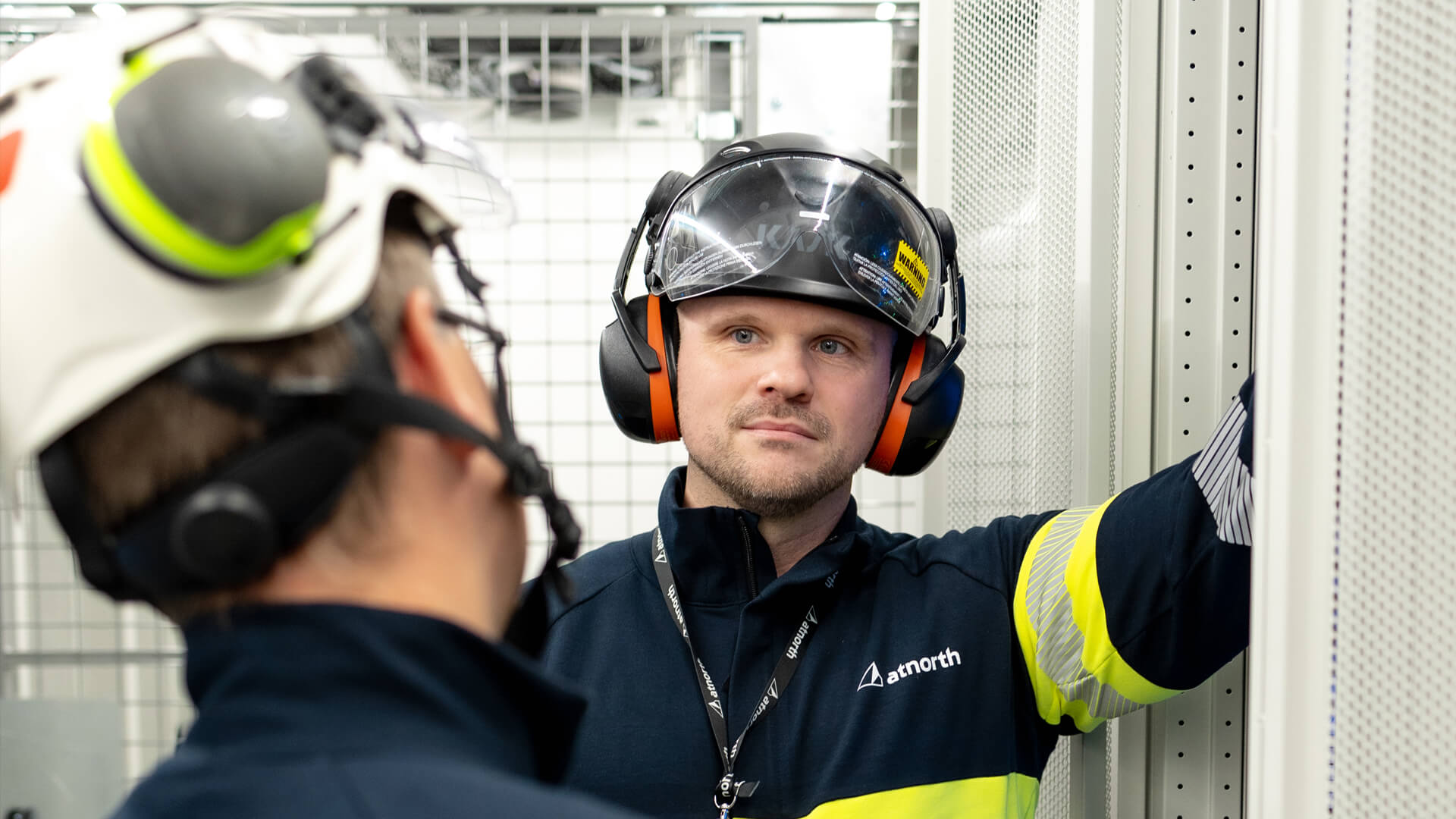 Two workers in safety gear converse near industrial equipment at the data center. One is wearing a protective helmet, earmuffs, and a high-visibility jacket.