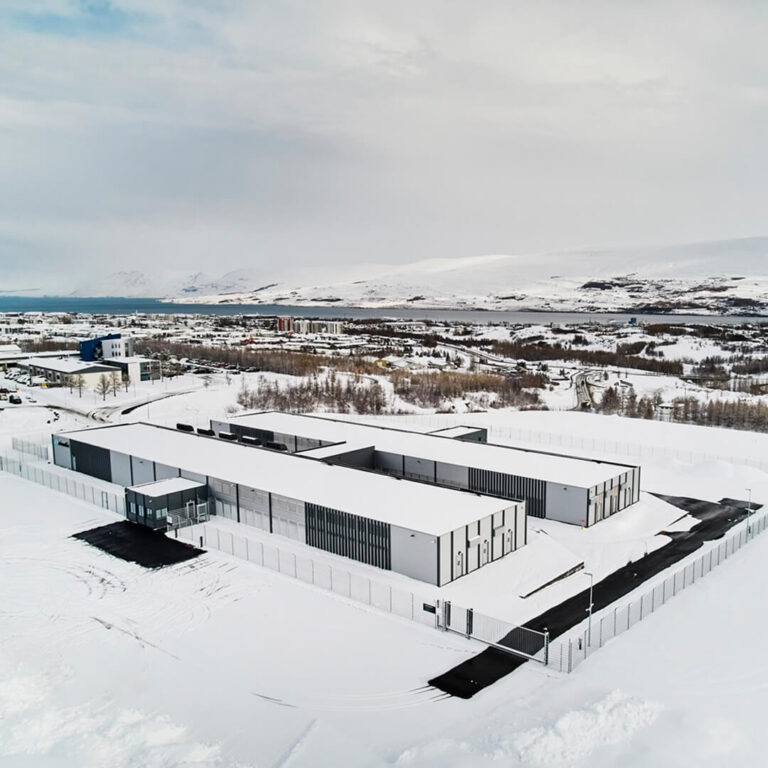 Aerial view of a sustainable data center complex in a snowy landscape, encircled by a fence, with distant mountains and a partly cloudy sky.