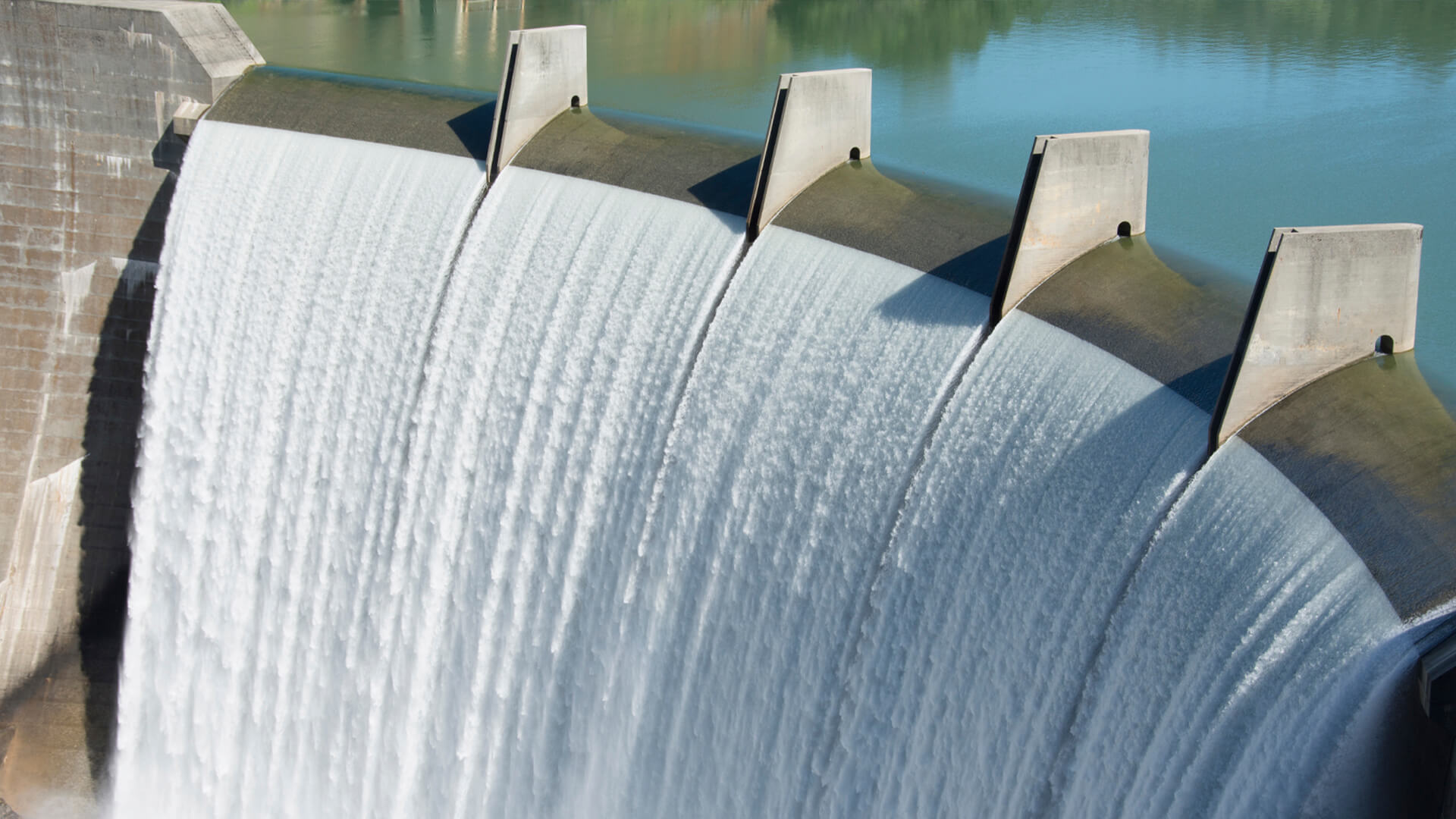 Water flows over the concrete spillway of a dam, creating a cascading effect reminiscent of cooling systems in a data center. In the background, tranquil waters and lush greenery complete the serene scene.
