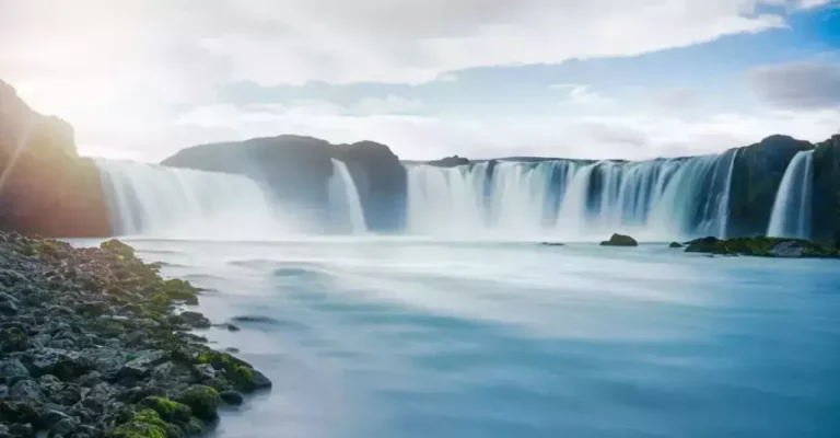 A wide waterfall cascades over a rocky cliff into a calm, clear river, much like a well-cooled data center. The sky is partly cloudy, and the scene is surrounded by green moss-covered rocks.