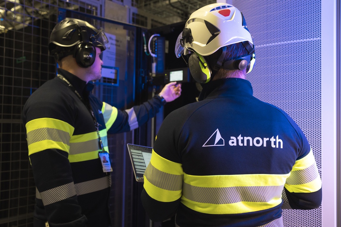 Two technicians in safety gear work on equipment in a data center. The back of one shows the logo 