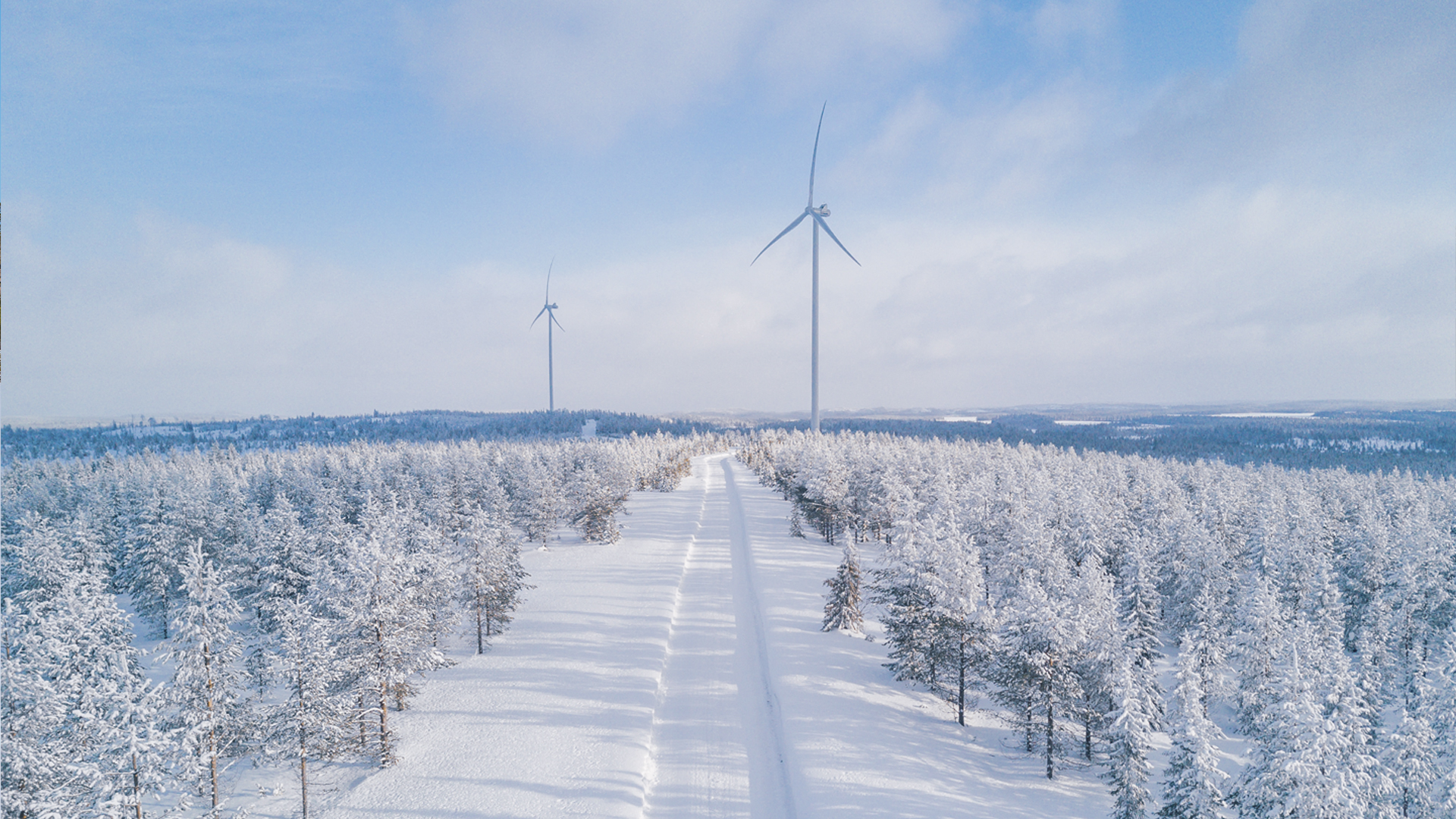 Snow-covered landscape with two wind turbines standing in a forest under a cloudy blue sky. A narrow road cuts through the trees.