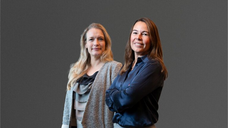 Two women stand side by side against a gray background, one with long blonde hair and a gray blazer, and the other with long brown hair and a dark shirt, both smiling.