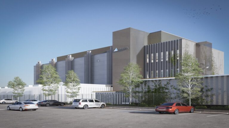 Modern industrial building with several parked cars in the foreground. Tall, minimalist design with trees nearby and a clear blue sky.