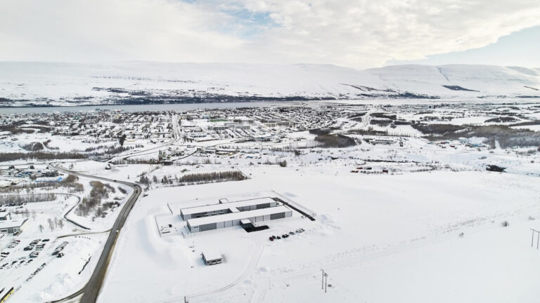 Aerial view of a snowy landscape with a small town, roads, and hills in the background under a partly cloudy sky.