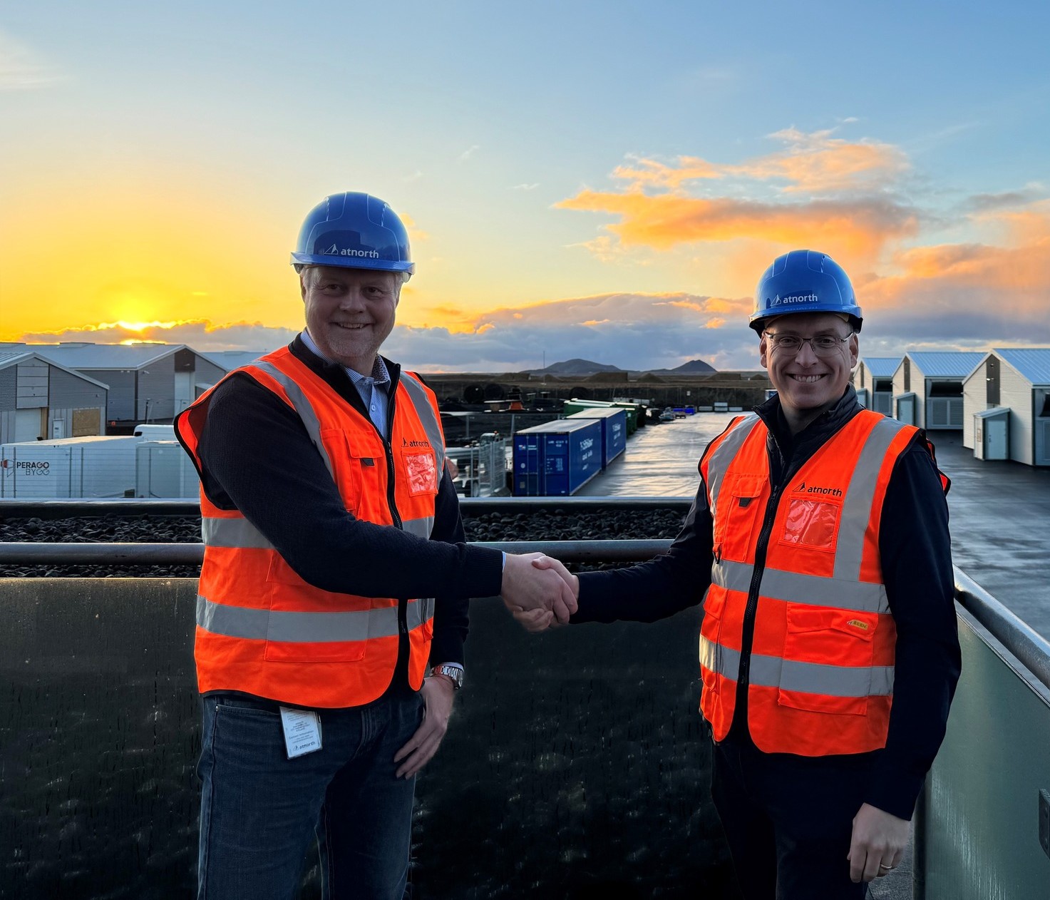 Two people in orange vests and blue hard hats shake hands at a construction site, with a sunset in the background.