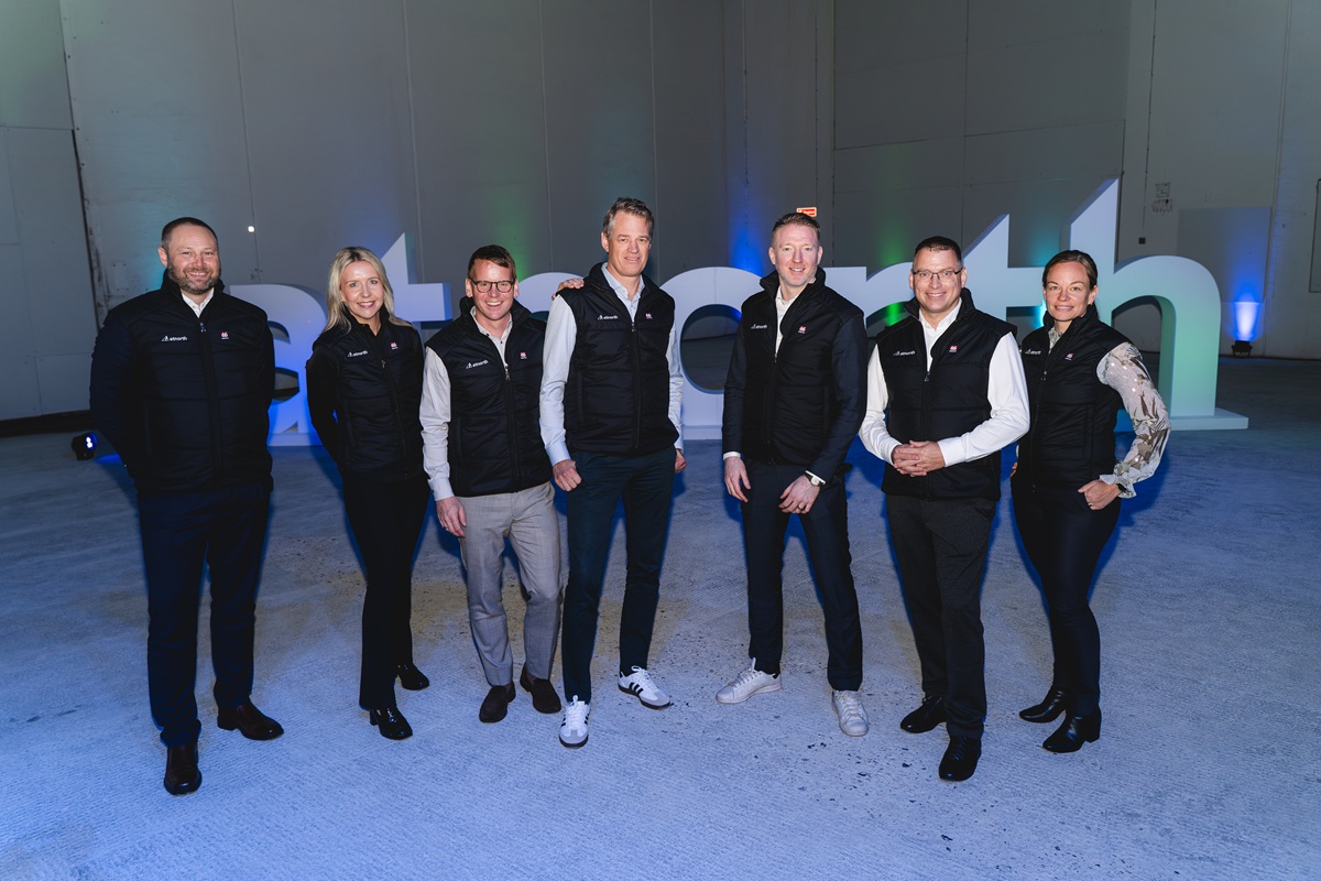 A group of seven people stands in a row indoors, all wearing black vests and business attire. The background features large, illuminated letters.