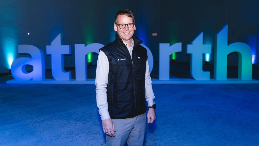 A person in a vest and glasses stands in front of large illuminated letters spelling "atnorth" in a blue-lit room.