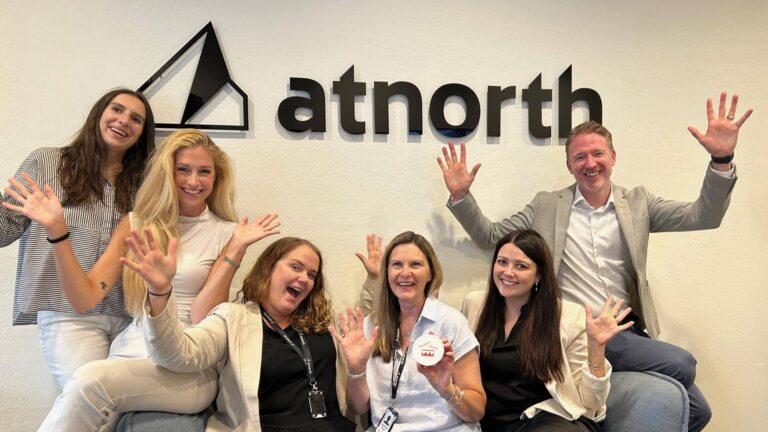 A group of six people smiling and waving at the camera while standing and sitting in front of an "atnorth" sign.