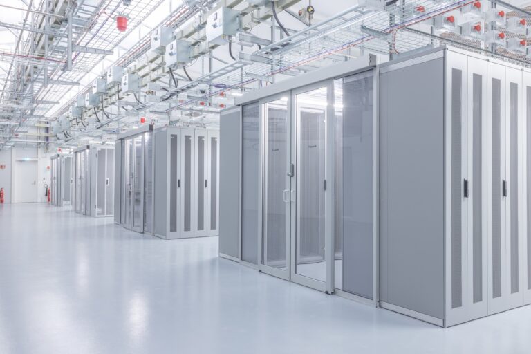 A modern data center with rows of server racks in a well-lit, clean environment. Glass doors and cable trays are visible above the racks.