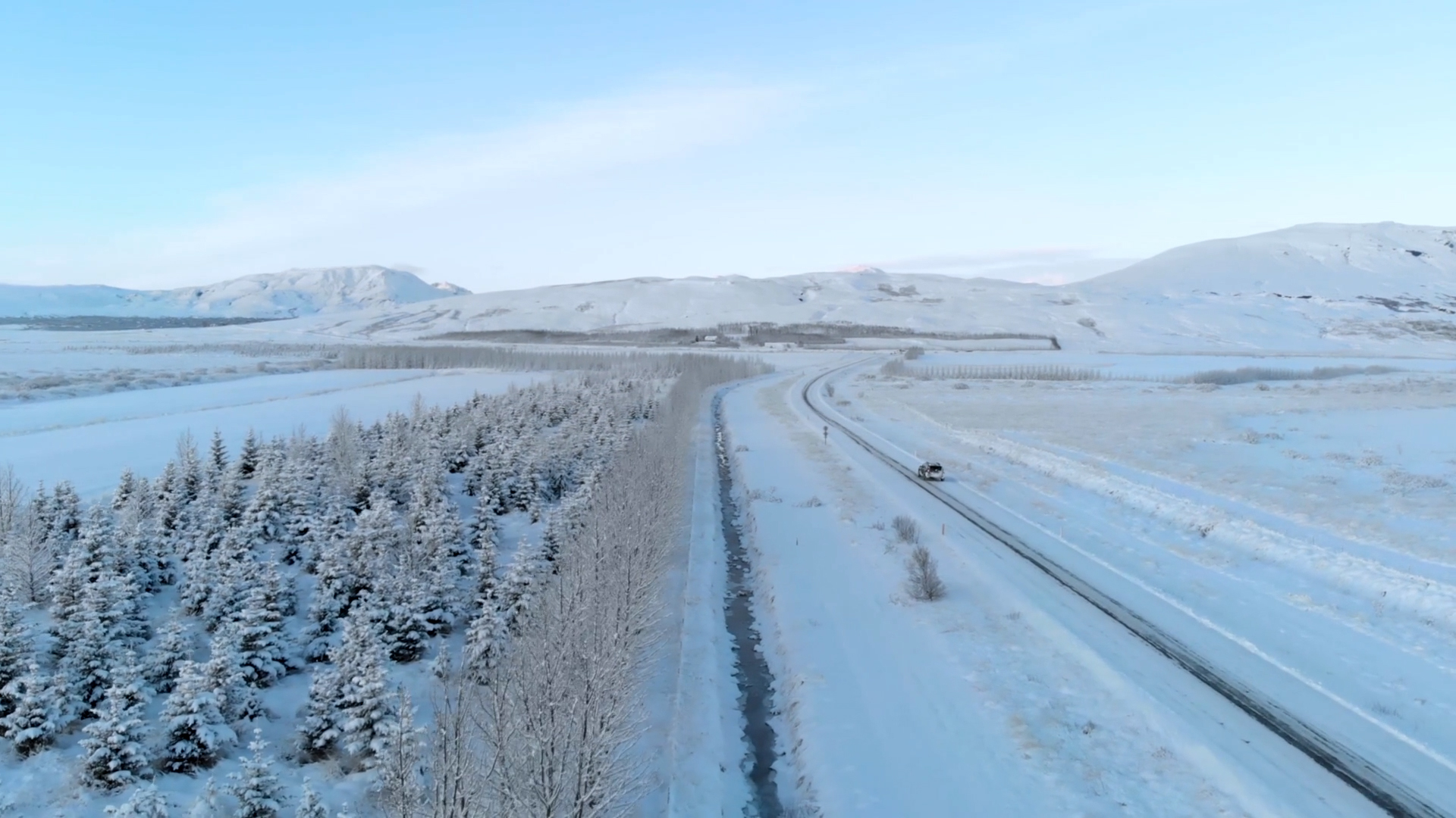 A snowy landscape with a road running through it, flanked by snow-covered trees on one side and open fields on the other. A vehicle is visible on the road under a clear, blue sky.