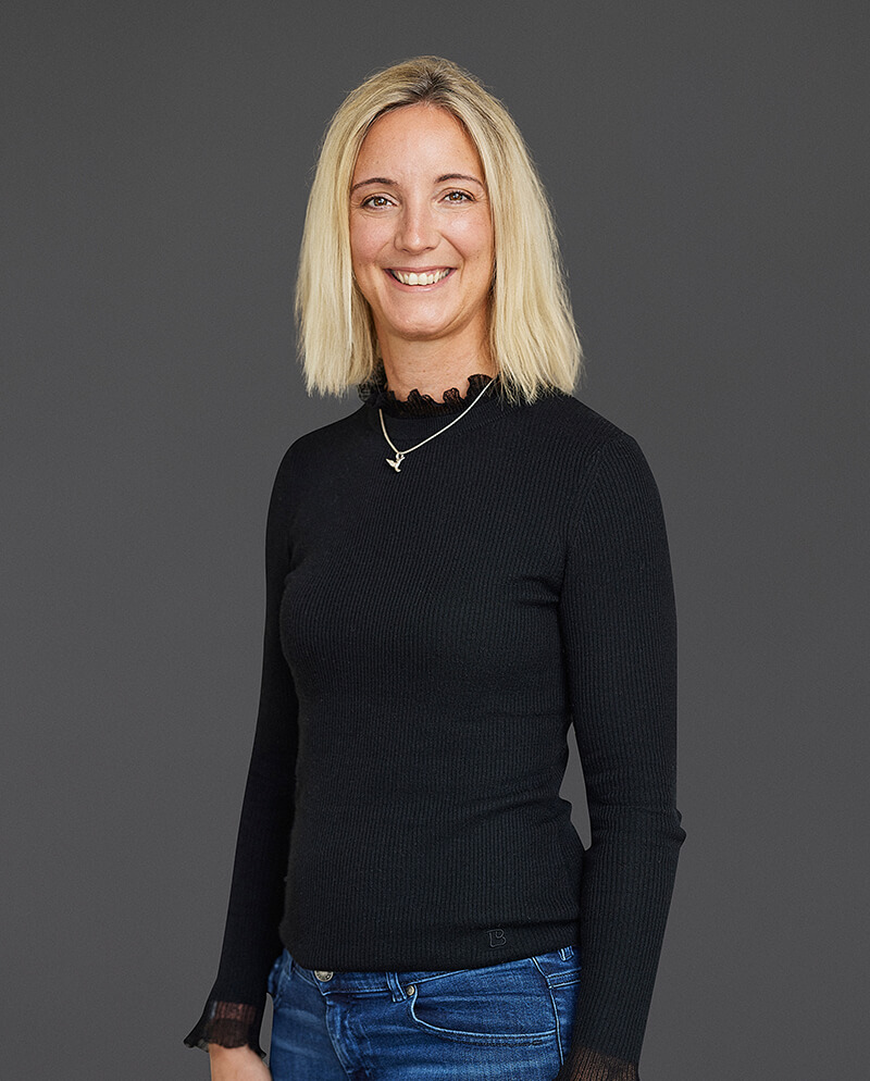A woman with shoulder-length blonde hair is smiling, wearing a black sweater and blue jeans, standing against a gray background.