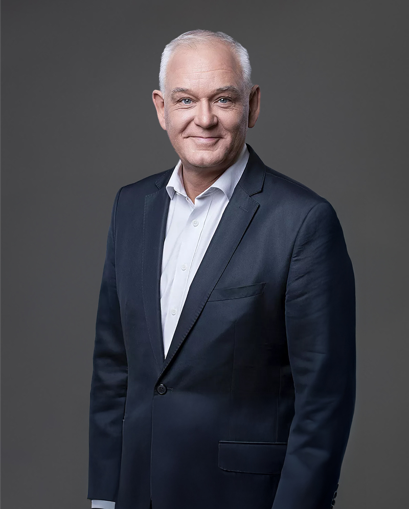 A bald man in a dark suit and white shirt stands against a gray background, smiling slightly.