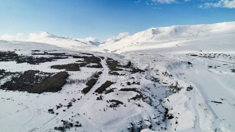 Aerial view of a snowy mountainous landscape with winding roads and scattered tree patches under a clear blue sky.
