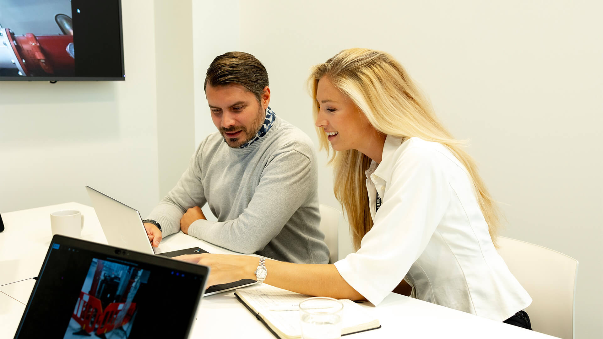 Two people sitting at a table working on laptops and a notebook in a bright office setting.