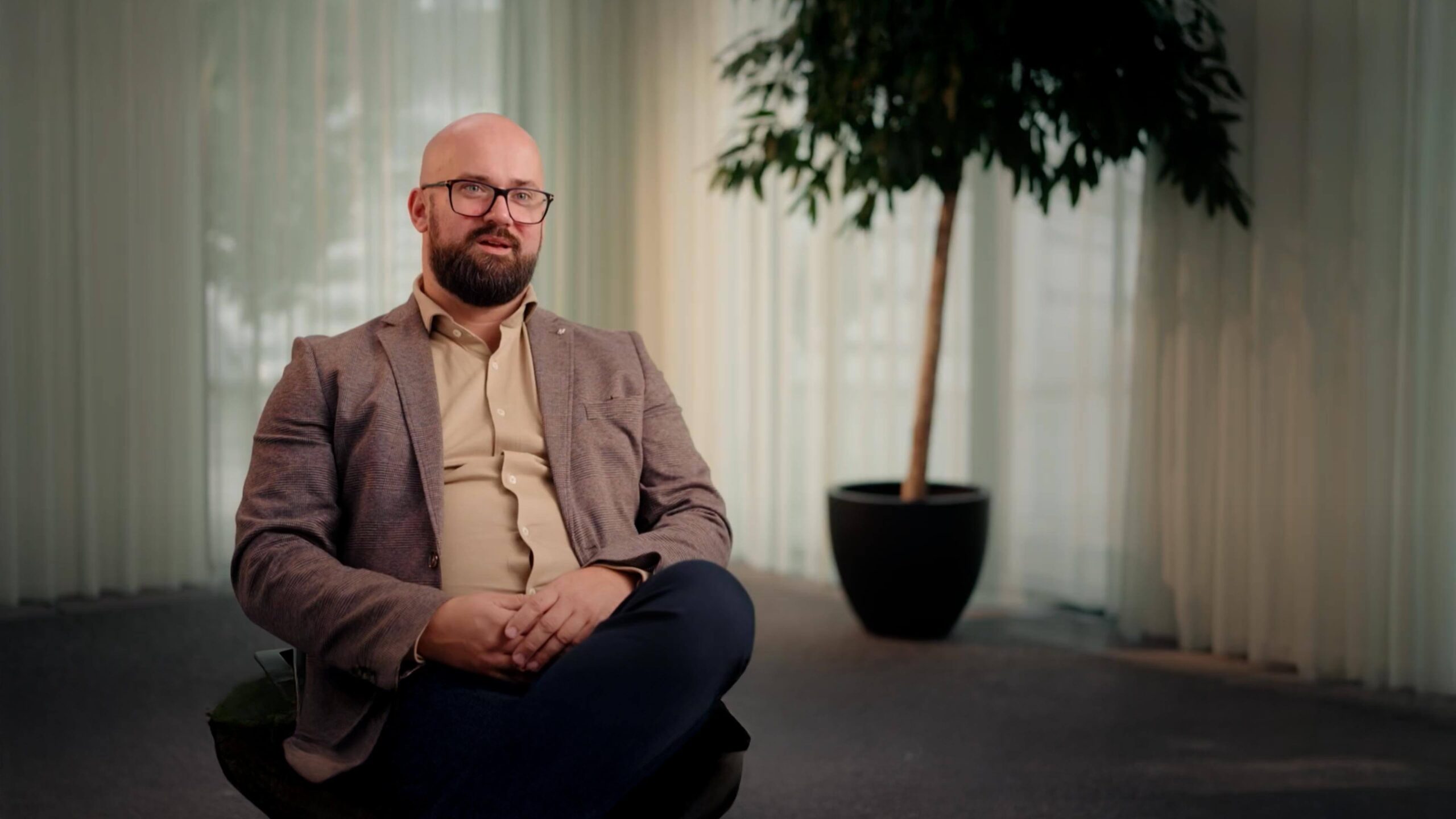 A person with a beard, glasses, and a blazer sits with crossed legs in a room with a large potted plant and sheer curtains.