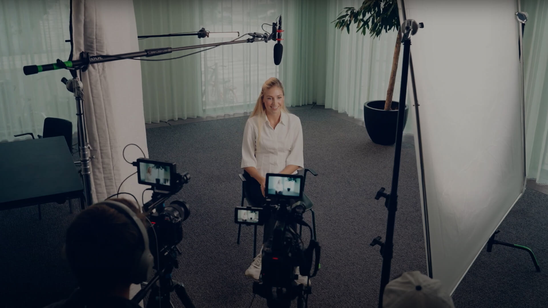 A person sits on a chair in a recording studio, surrounded by cameras, microphones, and lighting equipment.