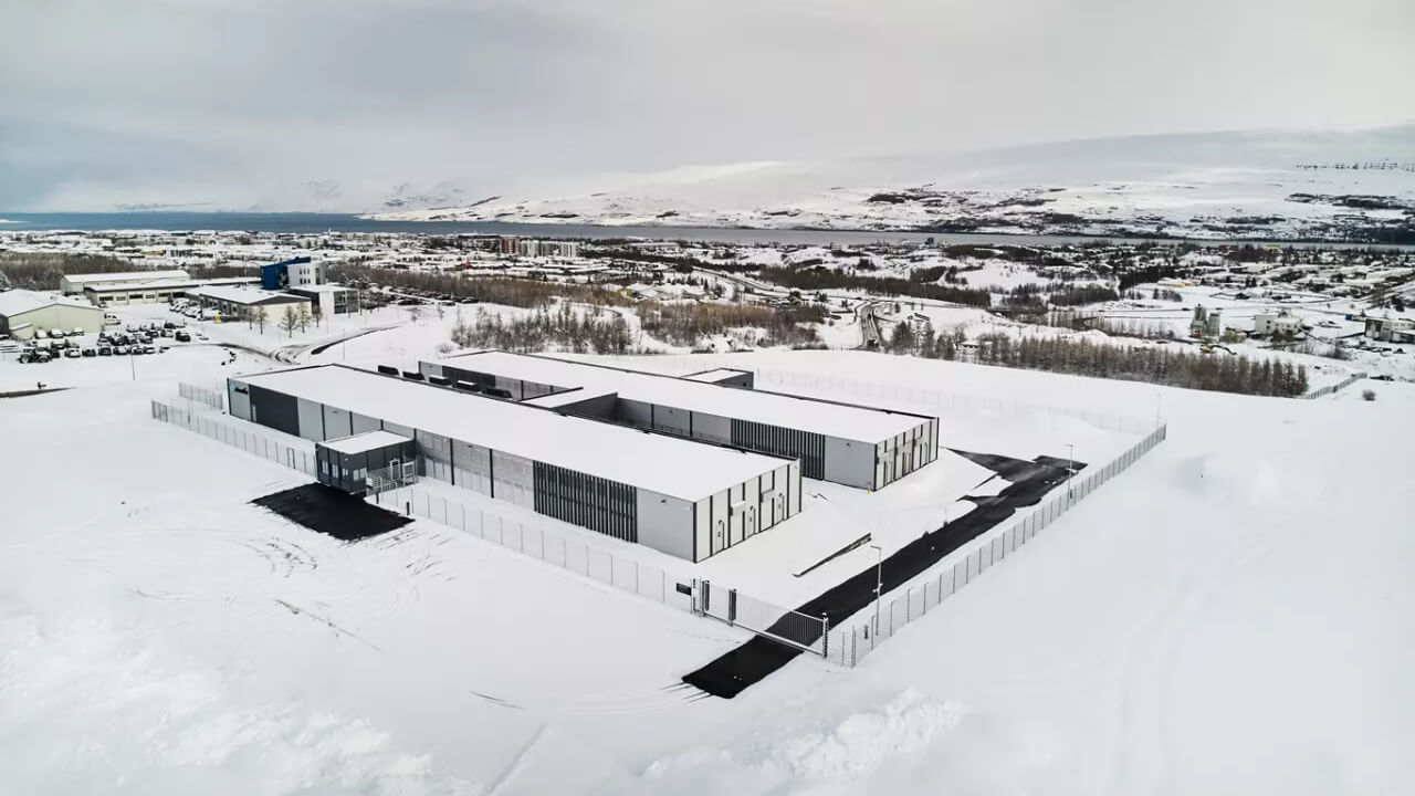 Aerial view of a large, modern building complex surrounded by snow, enclosed by a fence. Snowy landscape and distant town visible in the background.