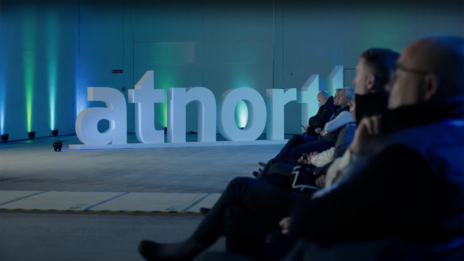 Several people seated in an auditorium facing a large "atnorth" sign on a stage with blue lighting.