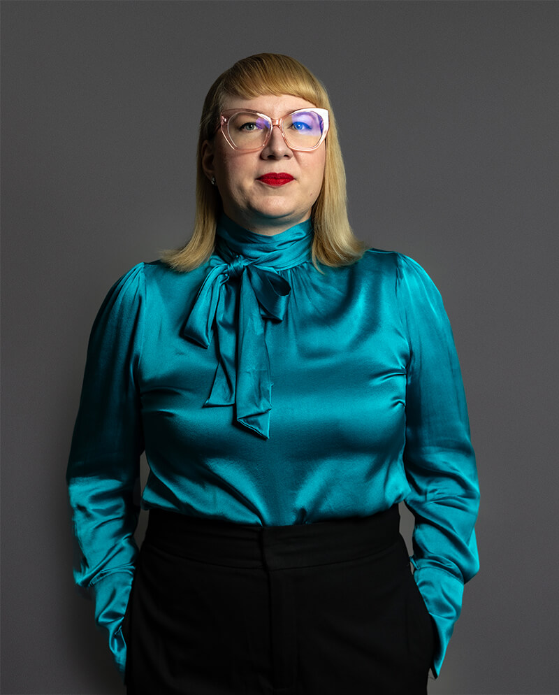 Person with glasses wearing a teal satin blouse with a bow, standing against a gray background.