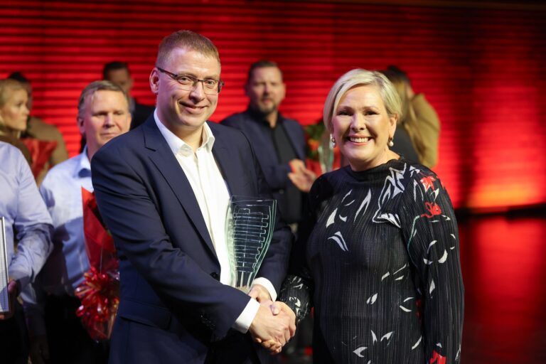 Two people in formal attire shake hands on a stage, with others in the background and a red backdrop. One person holds a glass award.
