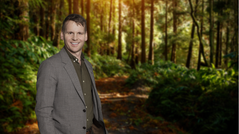 A man in a gray blazer and green shirt stands on a leafy forest path with sunlight filtering through the trees.