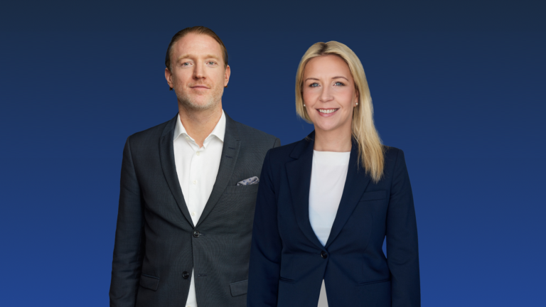 A man and woman in business attire stand in front of a blue gradient background. Both are looking forward.