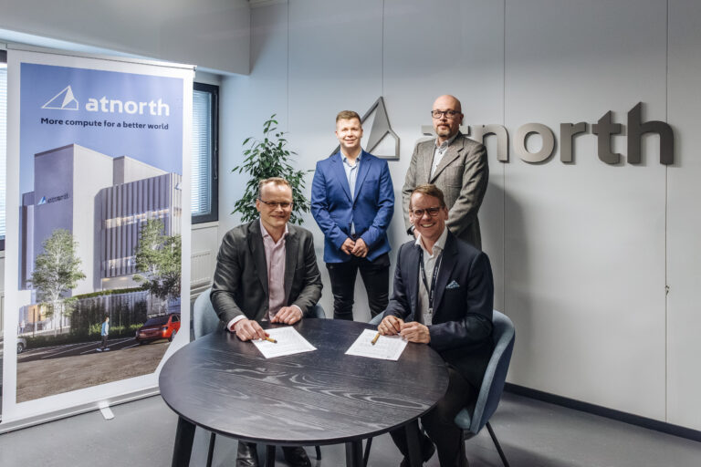 Four men at a signing ceremony in an office, with two seated at a table signing documents and two standing behind. A banner and wall sign display the "atnorth" logo.