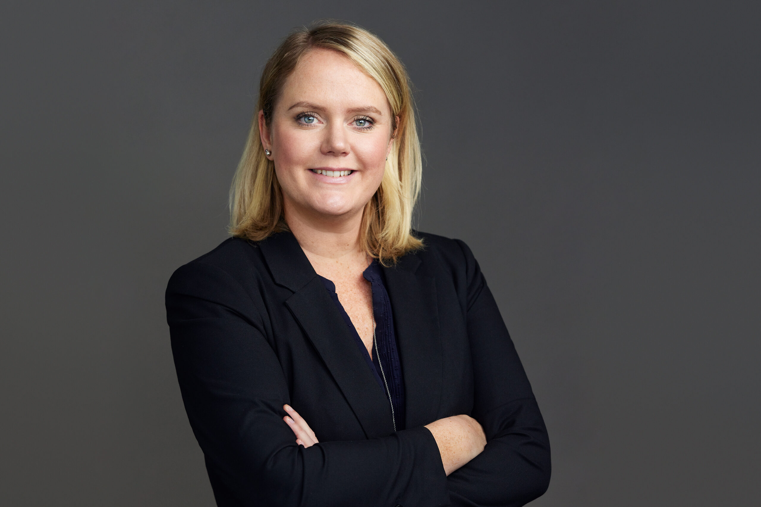 A woman with blonde hair in a black blazer stands against a plain gray background, arms crossed, and smiles at the camera.