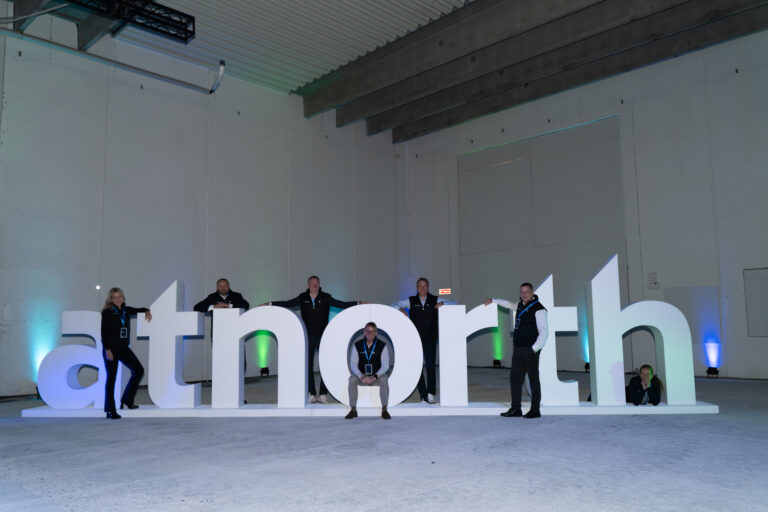 Six people pose around and on large white 3D letters spelling "atnorth" in an industrial indoor setting with subtle colored lighting.