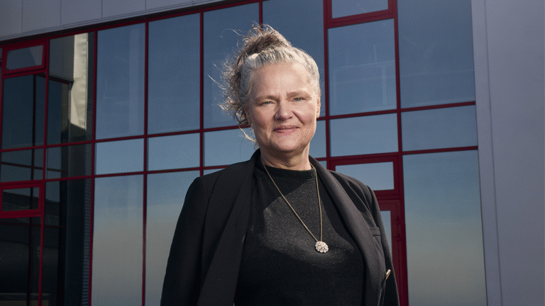 A woman with gray hair tied up, wearing a black outfit and pendant necklace, stands in front of a building with large reflective windows and red frames.
