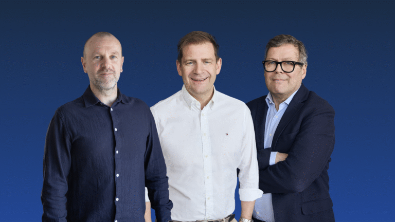 Three men stand side by side against a blue background. The man on the left wears a dark shirt, the middle man a white shirt, and the man on the right wears glasses and a dark jacket.