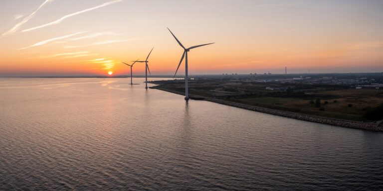 Three wind turbines stand along a shoreline at sunset, with calm water on one side and land on the other.