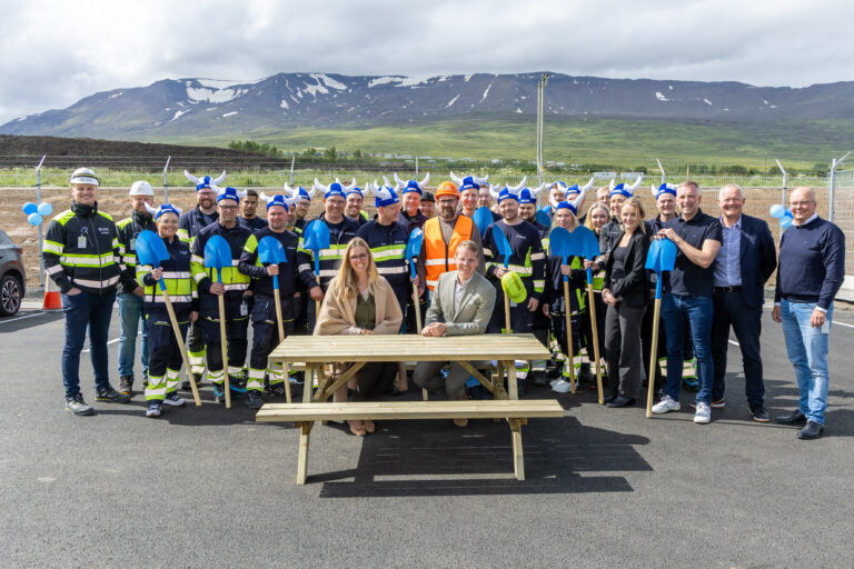 A group of people, some in work uniforms and Viking hats, pose outdoors with mountains in the background; three people sit at a picnic table in front.
