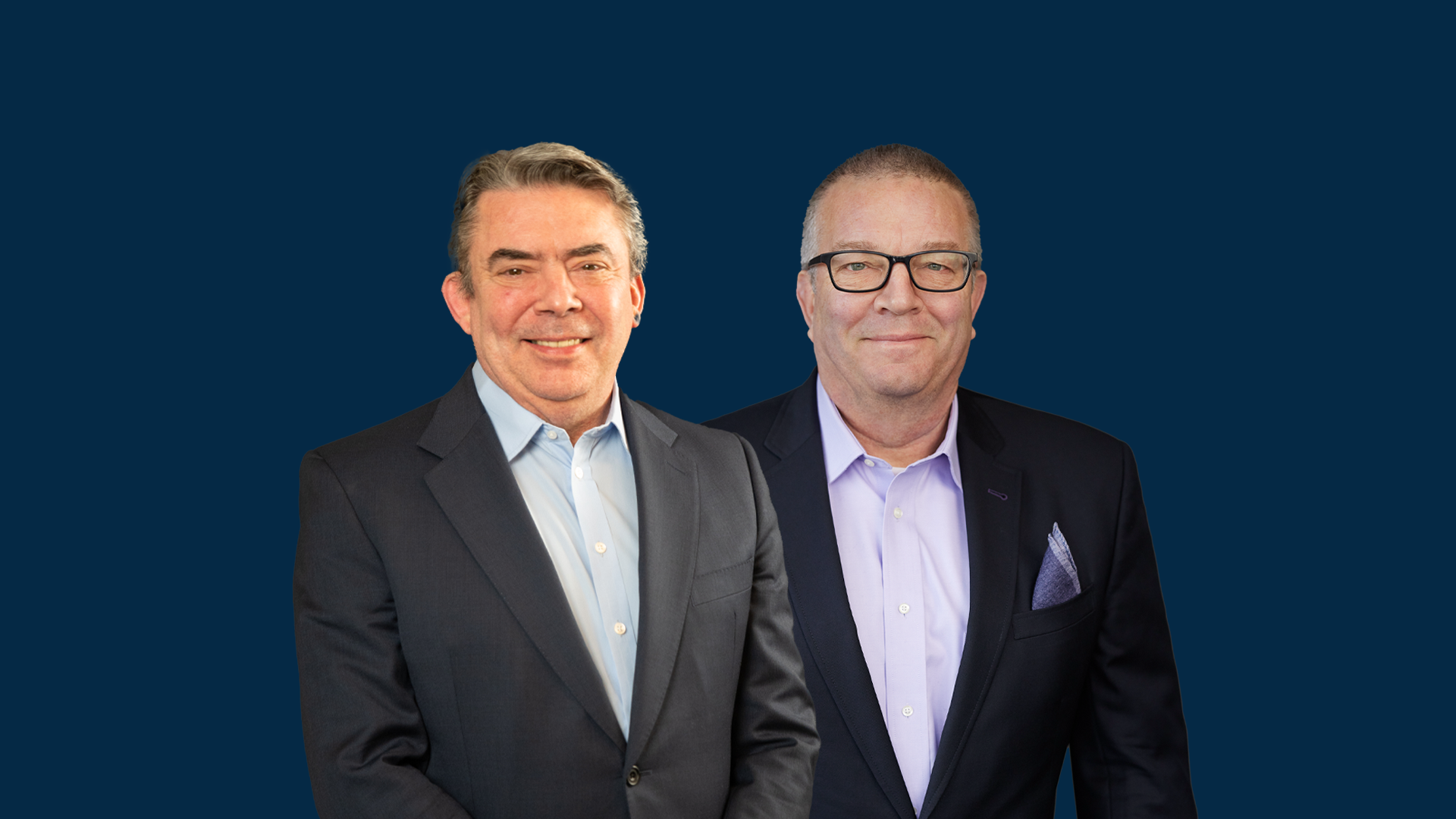 Two middle-aged men in business suits stand side by side against a solid dark blue background, both facing the camera and smiling.