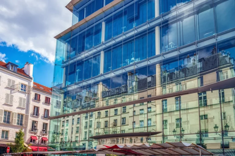 A modern glass building reflects the facade of traditional residential buildings and a partly cloudy sky in an urban setting.