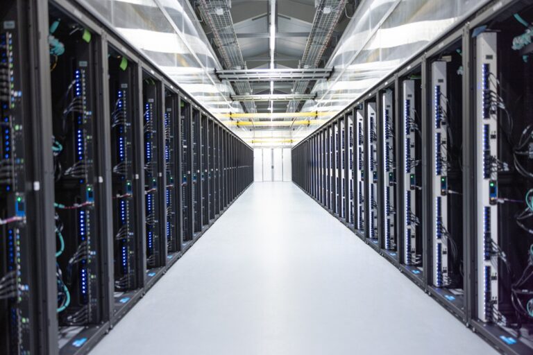 A data center aisle lined with server racks on both sides, showing network cables, illuminated indicator lights, and a brightly lit floor and ceiling.