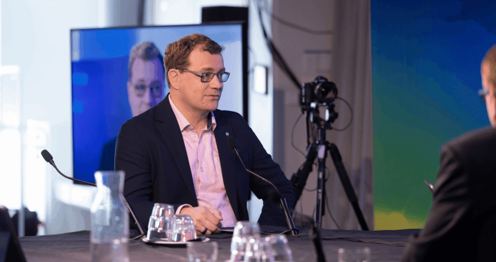 A man in a suit and glasses speaks into a microphone during an interview; his image is displayed on a screen in the background.